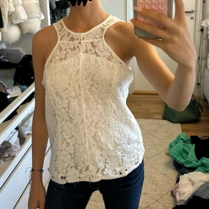 Vit spetsblus från Belair - Supersöt vit blus från Belair i spets med blommigt mönster. Ärmlös modell med hög halsringning och snygg passform. Perfekt för dig som gillar romantisk och feminin stil. Lätt och luftig, passar grymt till jeans eller kjol.