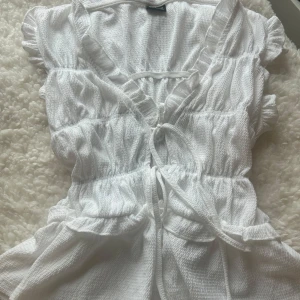 Vit volangblus med knyt från Gina Tricot - Supersöt vit blus från Gina Tricot i storlek XS. Toppen har volangdetaljer, v-ringning och knyt framtill. Den är ärmlös och har en lätt strukturerad bomullskänsla. Perfekt för en fräsch och somrig look!