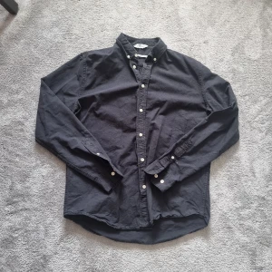 Svart skjorta från Samsøe Samsøe - Svart skjorta från Samsøe Samsøe i klassisk modell med vita knappar och button down-krage. Skjortan är långärmad och har en normal passform. Perfekt för dig som gillar stilrena och enkla plagg med snygg finish.