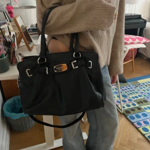 Svart axelväska från Michael Kors - Perfekt skolväska!Funkar till allt. As snygg svart axelväska från Michael Kors med silverdetaljer och metallskylt framtill. Väskan har både handtag och axelrem, perfekt för att bära på olika sätt. Tillverkad i skinn med stilrena spännen och klassisk form. Repor på metall brickan och och lite skadad vid ena handtaget.