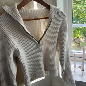 Vit ribbad stickad tröja med half zip - Superfräsch vit stickad tröja med ribbat mönster och halv dragkedja framtill. Tröjan har hög krage och långa ärmar, perfekt för lager på lager. Snygg och enkel att styla till jeans eller kjol.
