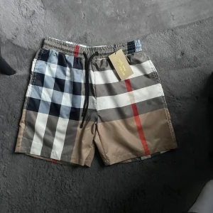 Rutig Burberry shorts med snörning - Snygga Burberry shorts med klassiskt rutigt mönster i beige, blått, vitt och rött. De har elastisk midja med svart snörning och är gjorda i lätt polyester, perfekt för sommaren. Stilen är loose och de har meshfoder på insidan för extra komfort.