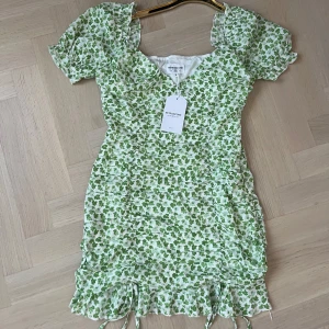 Grönblommig kort klänning Retro & Icone - Supersöt kort klänning från Retro&Icone med grönt blommigt mönster på vit botten. Puffiga korta ärmar, v-ringad hals och rynkade detaljer längs sidorna. Klänningen har dragkedja i sidan och volangkant nedtill. Perfekt för sommarens alla tillfällen!