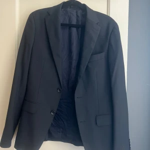 Svart kavaj These Glory Days slim fit - Stilren svart kavaj från These Glory Days i slim fit-modell. Klassisk design med två knappar framtill, tre fickor och snyggt foder. Perfekt för dig som gillar en smalare passform och skandinavisk stil. Passar bra om du är 170.