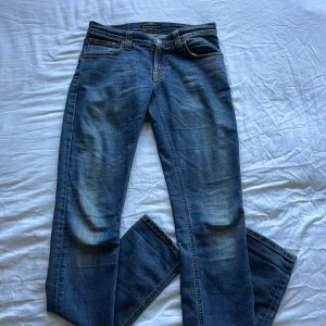 Nudie Jeans blå jeans W27 L34 - Snygga blå jeans från Nudie Jeans med klassisk femficksdesign och kontrastsömmar. Jeansen har raka ben och subtila slitningar framtill. Bakfickorna har Nudie Jeans karaktäristiska broderi. Tillverkade i bomull för en skön känsla.
