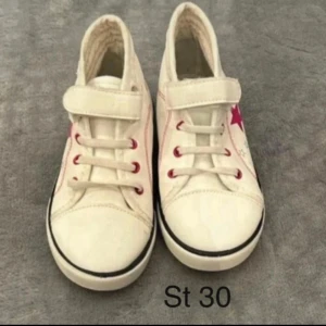 Vita sneakers med röd stjärna, stl 30 - Snygga vita sneakers i storlek 30 med röd stjärndetalj på sidan och röda öljetter. Skorna har både snörning och kardborreband för enkel påtagning. Rund tå och platt sula ger en klassisk look. Perfekta för dig som gillar stilrena och coola detaljer.