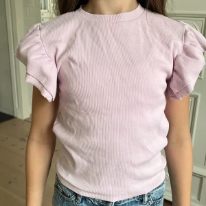 Ljusrosa ribbad topp med volangärm - Supersöt ljusrosa topp med korta volangärmar och ribbad struktur. Toppen har rund halsringning och är figurnära utan att vara tight. Perfekt för dig som gillar en feminin och trendig stil. Materialet känns mjukt och stretchigt.