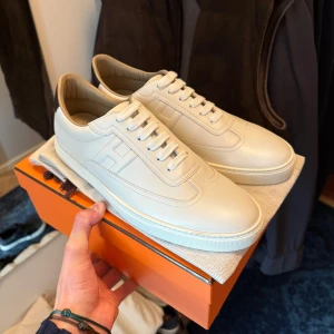 Vita Hermès sneakers i skinn - Exklusiva vita sneakers från Hermès i lyxigt skinn med diskret H-detalj på sidan och svart hälparti med Hermès-logga. Klassisk rund tå, snörning och platt sula. Perfekta för dig som vill ha en clean och stilren look med premiumkänsla.