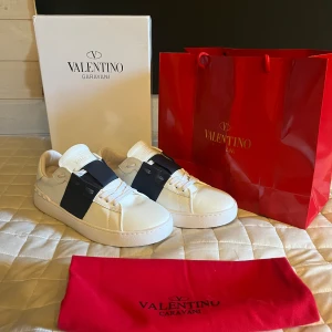 Valentino Garavani Open  - Säljer ett par Valentino Garavani Open i storlek 43 i vitt skinn med bred blå rem över vristen och coola nitar på hälen. Klassisk rund tå och snörning framtill. Kommer med originalkartong, dustbag och röd shoppingpåse. Perfekt för dig som gillar lyxiga detaljer.