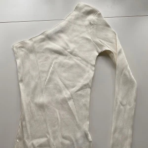 Vit oneshoulder topp från NA-KD - Säljer en vit ribbad oneshoulder topp från NA-KD i storlek XXS. Toppen har lång ärm på ena sidan och ärmlös på andra, vilket ger en cool asymmetrisk look. Materialet är mjukt och stretchigt, perfekt för att skapa en trendig outfit.