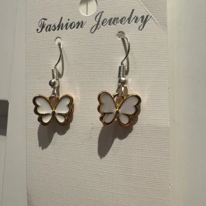 Vita fjärilsörhängen med guldkant - Superfina örhängen med vita fjärilar och guldfärgade kanter. Hängande modell med silvriga krokar, perfekt för dig som gillar söta detaljer och vill sticka ut lite extra. Fjärilarna har en stilren och lekfull design som passar till många olika looks.