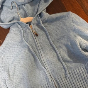 Ljusblå stickad hoodie med dragkedja - Säljer en ljusblå stickad hoodie med dragkedja framtill och huva. Tröjan har ribbade detaljer vid nederkant och ärmar, samt snörning vid huvan. Perfekt för chill dagar och enkel att matcha med jeans eller mjukisbyxor.