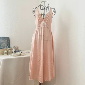 Vintage Olga klänning med spets (tag cut) - Supersöt klänning i aprikosfärg med spetdetaljet och djup v-ringning. Klänningen har broderade blommor framtill och baktill för att täcka några håll. Materialet är mjukt och faller fint, perfekt för sommarens alla tillfällen. Storlek: M (36-38). Dockans storlek: 34.