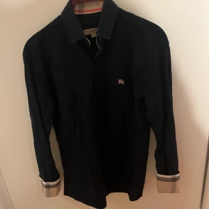 Svart Burberry skjorta med knappar - Snygg svart skjorta från Burberry London i storlek S. Klassisk design med långa ärmar och knappar framtill. Ikonisk Burberry-logga broderad på bröstet och typiska rutiga detaljer vid ärmsluten. Perfekt för dig som gillar stilrena plagg med lyxig touch.