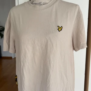 Beige t-shirt från Lyle & Scott - Snygg beige t-shirt från Lyle & Scott med klassisk rund hals och korta ärmar. På bröstet sitter den ikoniska gula fågel-loggan som ger en clean och stilren vibe. Tillverkad i mjuk bomull som känns skön mot huden. Perfekt för en chill och enkel look.