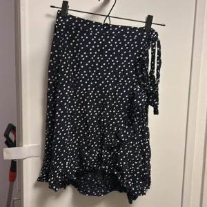 Mörkblå prickig omlottkjol Lindex - Supersöt mörkblå omlottkjol från Lindex med vita prickar och volangdetaljer. Kjolen har knytband i sidan och är perfekt för dig som gillar en lekfull och trendig stil. Lätt och skön i bomull, passar perfekt till sneakers eller sandaler.