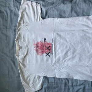 Off-White x KAWS vit t-shirt - Säljer en vit t-shirt från Off-White i samarbete med KAWS. Framsidan har en stor rosa och svart grafisk print med KAWS-figurer och Off-White-logga. Baksidan har ännu större print med text och illustrationer i rosa och svart. Klassisk passform och rund hals. Perfekt för dig som gillar streetwear. Ikke autentisk 