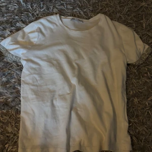 Vit t-shirt med glittriga ärmslut Zara - Vit t-shirt från Zara i storlek S med glittriga detaljer på ärmsluten. Klassisk rund hals och rak passform. Materialet är mjuk bomull som känns skönt mot huden. Perfekt för dig som gillar enkla plagg med en twist.