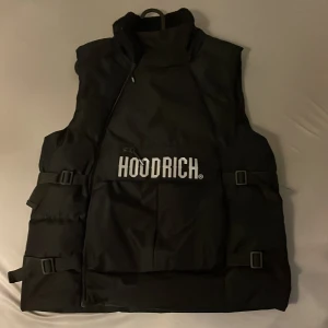 Svart dunväst från Hoodrich - Säljer en svart dunväst från Hoodrich med stor logga framtill. Västen har hög krage, justerbara spännen på sidorna och en stor ficka med dragkedja på magen. Perfekt för lager-på-lager och streetwear-stil.