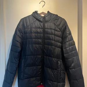 Svart pufferjacka med huva - Snygg svart varm pufferjacka med huva och röd insida. Jackan har dragkedja framtill, två fickor och quiltad design. Perfekt till höst och vinter 