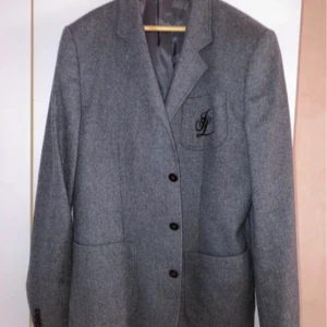 Grå kavaj från J.Lindeberg - Stilren grå kavaj från J.Lindeberg med klassisk passform. Kavajen har tre knappar framtill, två stora fickor och en bröstficka med broderad JL-logga. Tillverkad i ullblandning med slät fodring för en snygg och bekväm look.