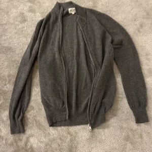 Grå kofta i kashmir från Soft Goat - Säljer en stilren grå kofta från Soft Goat i 100% kashmir. Koftan har långärmad design, ribbade muddar och dragkedja framtill. Perfekt för dig som gillar mjuka och lyxiga material. Enkel att matcha med jeans eller chinos.