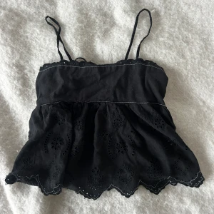 Svart broderad linnetopp med spets - Supersöt svart linnetopp med tunna axelband och broderade blommor. Toppen har volangig nederkant med spetsdetaljer och knytning i ryggen. Perfekt för varma dagar när du vill ha en chill och snygg look.