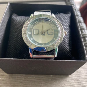 Nya Silverfärgad DqG klocka med meshband  - Nya Stilren klocka från DqG i silver med glittrande stenar runt urtavlan. Klockan har ett klassiskt runt ur och ett snyggt mesharmband i metall. Stor DqG-logga på urtavlan och tydliga siffror. Perfekt accessoar för dig som gillar att sticka ut.