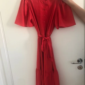 Röd långklänning med knytband (kostat 700kr innan) - Snygg röd långklänning med korta ärmar och knytband i midjan. Klänningen har knappar framtill och ett flowigt fall med volang nedtill. Perfekt för dig som gillar färg och vill ha en bekväm och stilren look.