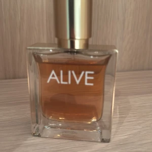 Hugo Boss Alive Eau de Parfum - Alive från Hugo Boss är en elegant parfym i en stilren, fyrkantig glasflaska med guldig kork. Doften har en varm bärnstensfärgad ton och flaskan har tydlig vit text framtill. Perfekt för dig som gillar lyxiga och moderna dofter.