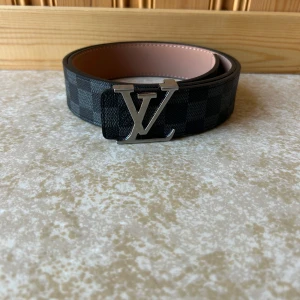 L-V Damier skärp med L-V-spänne - Snyggt L-Vbälte i klassiskt svart och grått Damier-mönster med ikoniskt L-V-spänne i silver. Bältet är gjort i skinn med ljusbrun insida och har exklusiva detaljer. Perfekt accessoar för att lyfta din outfit med lyxig känsla.