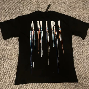 Svart AMIRI t-shirt med färgstänk - Svart t-shirt från AMIRI med stort vitt logotryck på ryggen och coola färgstänk i blått, orange och vitt som rinner ner. Klassisk rund hals och korta ärmar. Perfekt statement-plagg för dig som gillar streetstyle och unika detaljer.