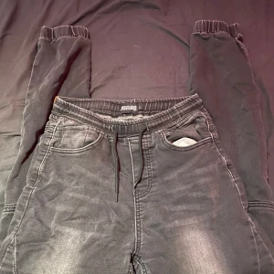 Svarta bikerinspirerade byxor Denimday - Svarta bikerinspirerade byxor från Denimday med elastisk midja och snörning. Byxorna har ribbade detaljer över knäna och muddar vid bensluten. Snygg tvättad look och flera fickor fram och baktill. Perfekta för en cool streetstil.