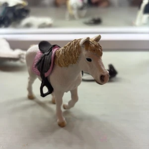 Schleich häst med rosa sadel och brunt tränns - Ingen bok syns på bilderna, utan det är en leksakshäst med sadel och träns. Om du letar efter böcker om hästar, ridning eller djur, vänligen ladda upp bilder på en bok.