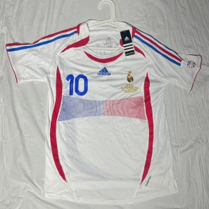 Frankrike Zidane VM 2006 tröja Adidas - Säljer en officiell Frankrike fotbollströja från VM 2006 med Zidane och nummer 10 på ryggen. Tröjan är vit med blå och röda detaljer, kortärmad och har Adidas-logga på bröstet. Materialet är lätt och andas, perfekt för match eller träning.