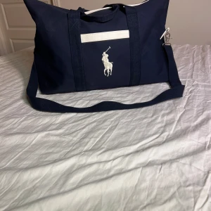 Blå weekendbag från Polo Ralph Lauren - Snygg marinblå weekendbag från Polo Ralph Lauren med stort vitt broderat logotyp framtill, vita detaljer och brunt skinn i botten. Väskan har både handtag och justerbar axelrem, perfekt för resor eller gymmet. Materialet är tålig bomullscanvas och skinn.