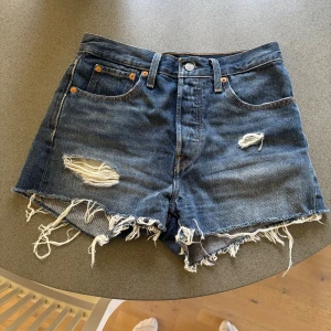 Levi's blå jeansshorts - Säljer ett par blå jeansshorts från Levi's med råa, fransiga kanter och slitna detaljer framtill. Märkespatch baktill och röd Levi's-tag på bakfickan. 
