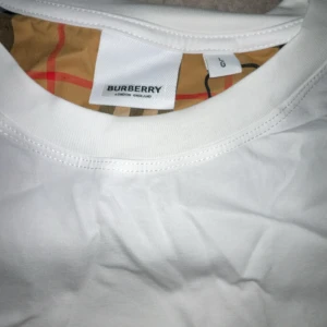 Vit Burberry t-shirt med rutiga ärmar - Snygg vit t-shirt från Burberry i storlek L. Klassisk rund hals och korta ärmar med Burberrys ikoniska beige, röda och svarta rutmönster. Tillverkad i mjuk bomull som känns skön mot huden. Perfekt för dig som gillar stilrena plagg med lyxig touch.