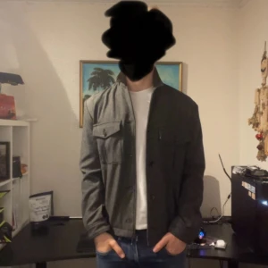 Grå overshirt från H&M, storlek M - Snygg grå overshirt från H&M i storlek M. Jackan har raka linjer, två bröstfickor med lock och knappar framtill. Perfekt att slänga över en t-shirt för en clean och stilren look. Materialet känns mjukt och passar bra till både jeans och chinos.