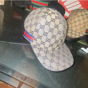 Gucci keps med GG-mönster och röd/blå detalj - Snygg Gucci keps i beige med klassiskt GG-monogram över hela kepsen. Har en röd och blå rand på sidan och justerbar rem baktill. Tillverkad i bomullsmix med marinblått innerfoder. Perfekt för dig som vill sticka ut med en lyxig accessoar.