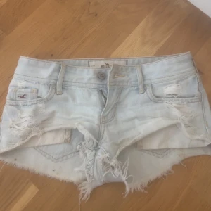 Ljusblå slitna jeansshorts Hollister - Säljer ett par ljusblå jeansshorts från Hollister med råa kanter och slitningar framtill och baktill. Klassisk femficksmodell med låg midja och dragkedja. Perfekta för sommaren och ger en avslappnad vibe.