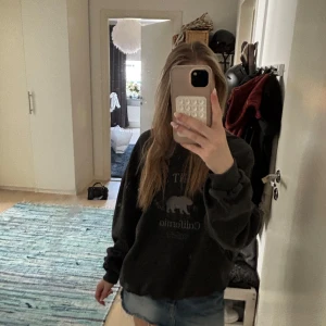 Svart sweatshirt Belmont Shore California - Svart sweatshirt med tryck 'Belmont Shore California' och björn framtill. Tröjan har rund hals, ribbade muddar och en loose fit. Perfekt för en chill och avslappnad stil. Materialet känns mjukt och skönt mot huden.