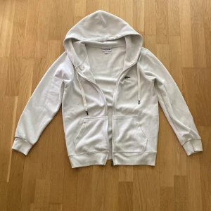 Lacoste hoodie - Vit Lacoste hoodie med dragkedja framtill, huva med snörning och två stora fickor. Klassisk och enkel design, stilren. Långärmad och mjuk, väldigt skön.