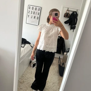 Vit broderad blus med volangärm - Superfin vit blus med broderade blommor och vågiga spetsdetaljer. Blusen har korta volangärmar, knappar framtill och en söt, rynkad halsringning. Perfekt för en fräsch och somrig look!