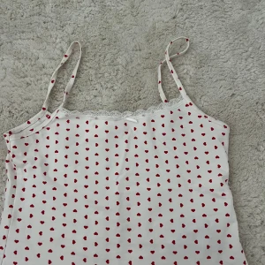 Vit pyjamaslinne med röda hjärtan Gina Tricot - Supersöt vit pyjamas linnetopp från Gina Tricot med röda hjärtan över hela tyget. Spaghettiband, spetskant upptill och en liten rosett framtill ger en gullig vibe. Perfekt för dig som gillar romantiska detaljer och vill ha en lätt pyjamas topp till sommaren.