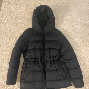 Svart pufferjacka från Everest - Svart pufferjacka från Everest med huva och dragsko i midjan. Jackan har långa ärmar, dragkedja framtill och två fickor. Snygga grå detaljer med Everest-logga på sidorna. Perfekt för kalla dagar när du vill hålla dig varm och stilren. Ny pris 999kr, pris går att diskutera 