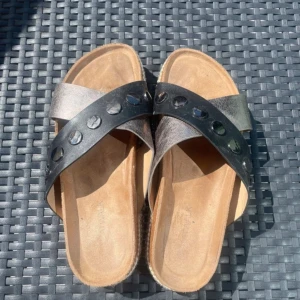 Silvriga sandaler med nitar och platåsula - Snygga sandaler med korsade remmar i metallic silver och svart, dekorerade med stora runda nitar. De har en bekväm plattformsula med flätad detalj runt kanten. Perfekta för att lyfta din sommarstil med en cool touch.