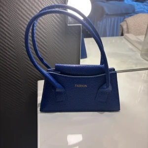 Blå minihandväska med guldtext - Snygg blå minihandväska i skinnimitation med två handtag och guldig text framtill. Väskan har en stilren, rektangulär form och stängs med magnetknapp. Perfekt för att bära det viktigaste och ge outfiten en pop av färg💓💓