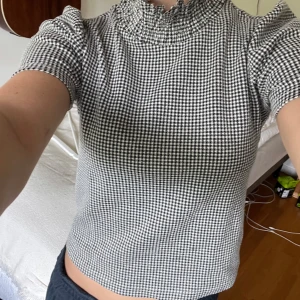 Svartvit rutig blus med puffärm - Säljer en svartvit rutig blus med korta puffärmar och hög smockad krage från Zara. Blusen har en cool vintagevibe och är gjord i mjukt, lätt material som känns skönt mot huden. Perfekt för dig som gillar att sticka ut med mönster och detaljer.