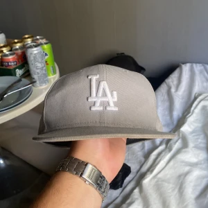 Grå LA keps från New Era 59FIFTY - Säljer en stilren grå keps från New Era 59FIFTY med LA-logga broderad i vitt framtill och MLB-logga bak. Kepsen har en klassisk rak skärm och är tillverkad i 100% polyester. Perfekt för dig som gillar streetwear och sportiga vibes.